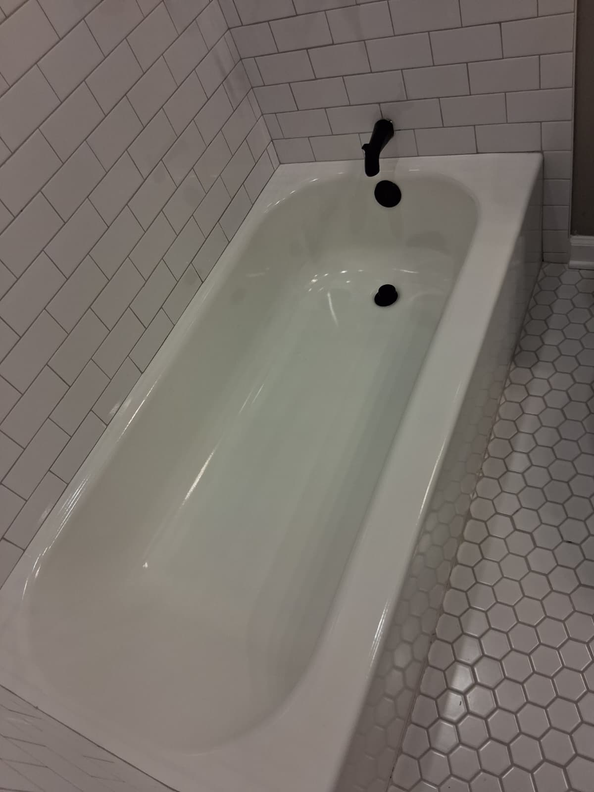 White bathtub with black fixtures, freshly cleaned and free of residue, surrounded by white subway tile walls and light hexagon tile flooring, representing a professionally cleaned bathroom.